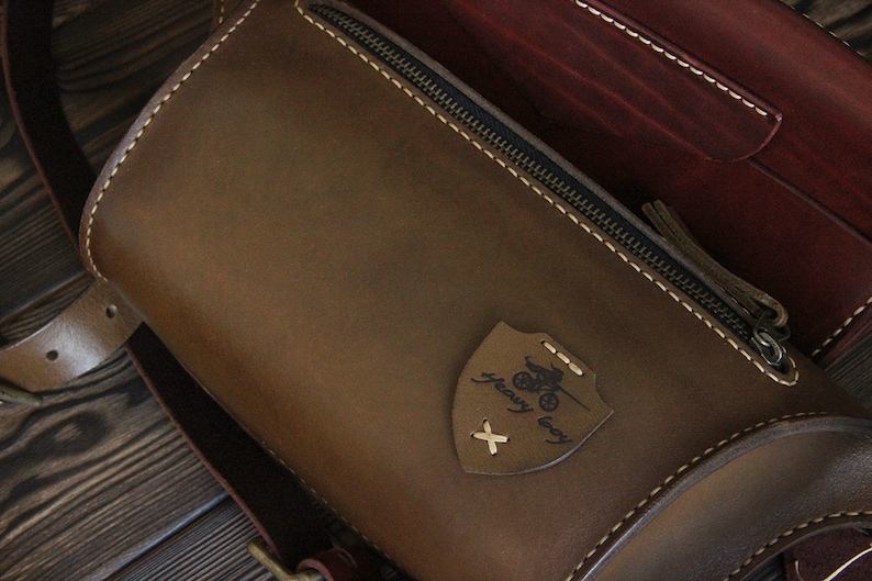 May include: A brown leather bag with a zipper closure and a small leather patch with the text "heavy duty" and a motorcycle graphic. The bag has a strap and is made of high-quality leather.