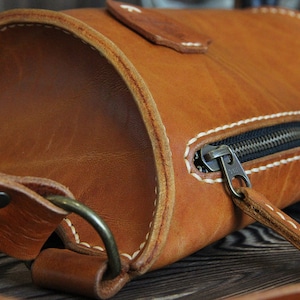 May include: A brown leather crossbody bag with a zipper closure and a white stitch detail. The bag has a strap and a metal buckle.