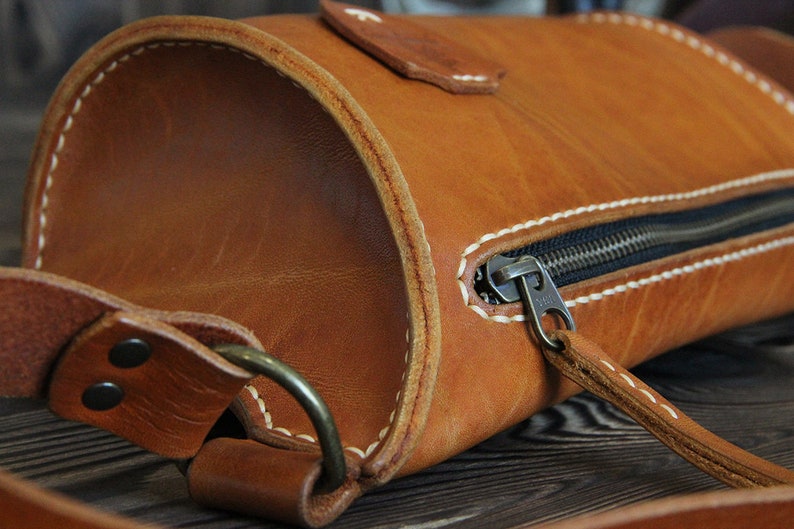 May include: A brown leather crossbody bag with a zipper closure and a white stitch detail. The bag has a strap and a metal buckle.