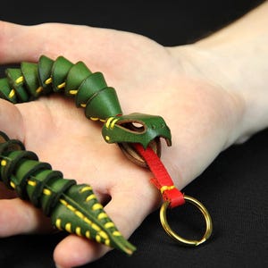 Op de afbeelding: Een groene leren slangen sleutelhanger met gele stiksels en een rode leren tong. De slang is opgerold en heeft een messing sleutelring.