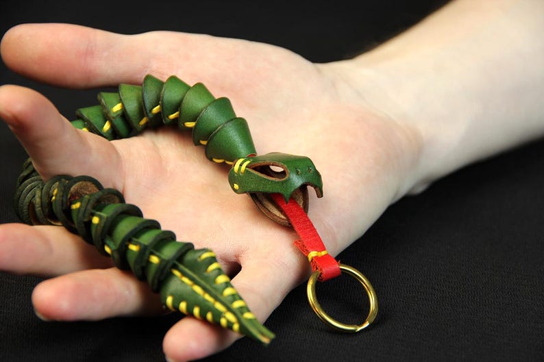 Op de afbeelding: Een groene leren slangen sleutelhanger met gele stiksels en een rode leren tong. De slang is opgerold en heeft een messing sleutelring.