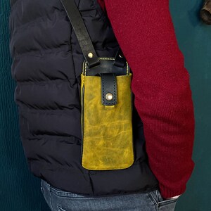 May include: A yellow leather phone case with a black strap, worn over the shoulder. The case has a flap that closes with a snap.