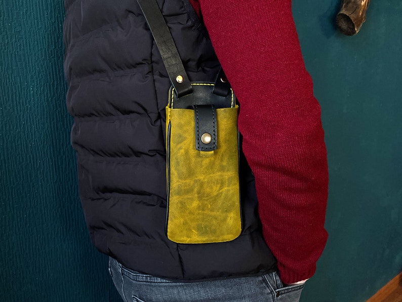 May include: A yellow leather phone case with a black strap, worn over the shoulder. The case has a flap that closes with a snap.