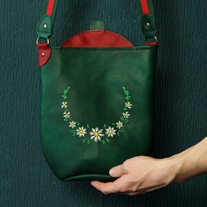 May include: A green leather crossbody bag with a red flap and a white daisy embroidery design.