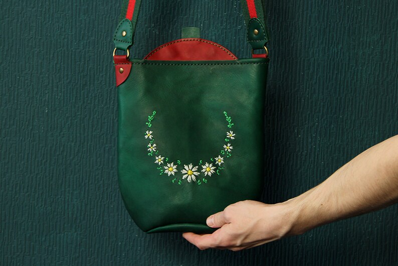 May include: A green leather crossbody bag with a red flap and a white daisy embroidery design.