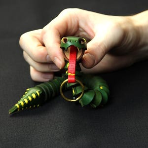 Op de afbeelding: Een groene leren slangen sleutelhanger met een rode tong en gele accenten. De slang is om een gouden metalen ring gewikkeld.