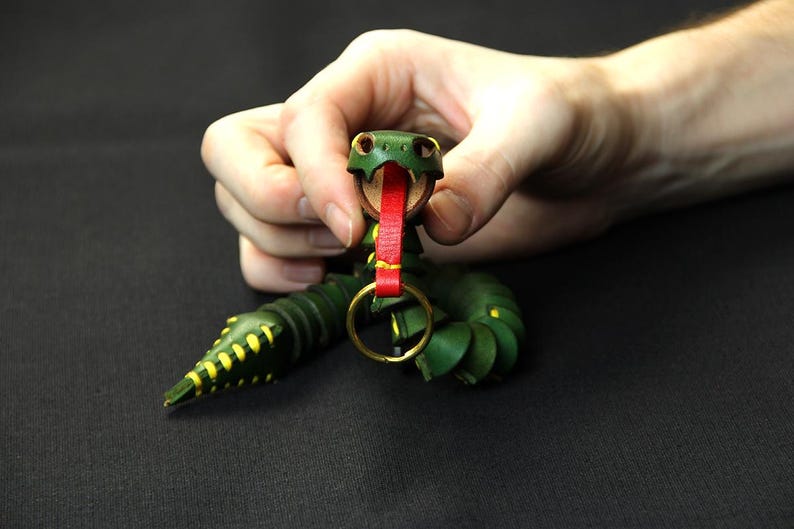 Op de afbeelding: Een groene leren slangen sleutelhanger met een rode tong en gele accenten. De slang is om een gouden metalen ring gewikkeld.
