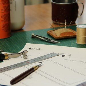 May include: A green cutting mat with a ruler, a leather awl, a metal tool, a small metal compass, a spool of thread, a leather needle holder, and a piece of paper with a leather pattern on it.