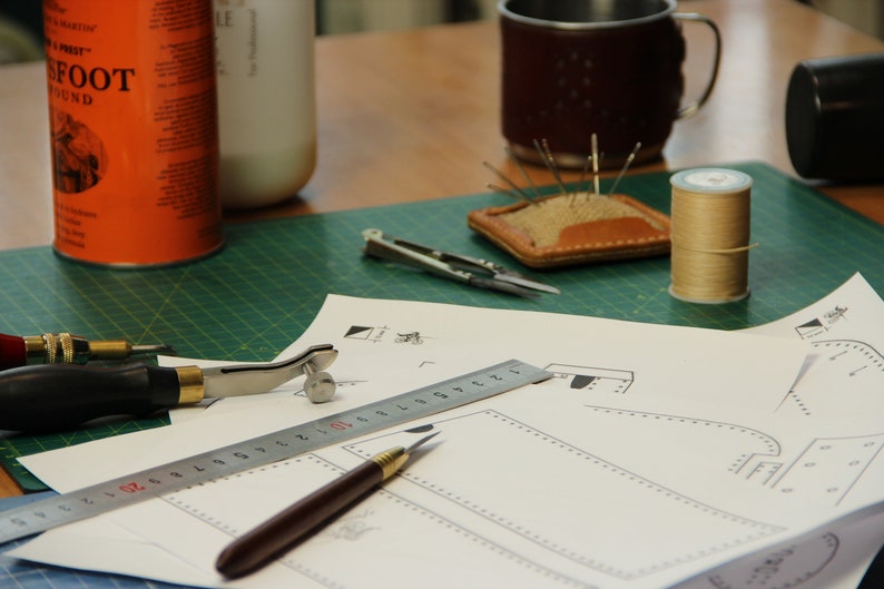 May include: A green cutting mat with a ruler, a leather awl, a metal tool, a small metal compass, a spool of thread, a leather needle holder, and a piece of paper with a leather pattern on it.