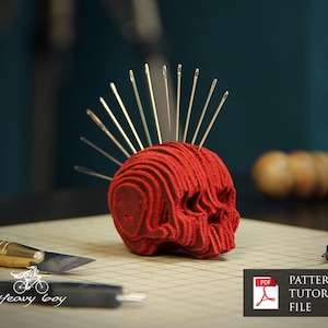 Puede incluir: Un cojín de alfileres rojo en forma de calavera, con agujas insertadas para parecer un mohawk. La imagen incluye un cuchillo artesanal, una tenaza de cuero y un archivo de patrón PDF, lo que sugiere un proyecto de artesanía en cuero DIY.