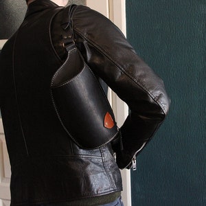 May include: A black leather crossbody bag with a white stitch detail and a brown leather accent. The bag is worn over the shoulder of a person wearing a black leather jacket.