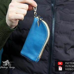 May include: A blue leather key pouch with a gold zipper and white stitching. The pouch is held by a silver clasp. The background includes a black vest and a green sweater. The pouch has a small leather tab.