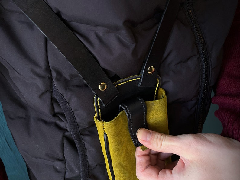 May include: A yellow leather phone case with black leather straps and gold accents. The case is attached to a black leather strap that goes over the shoulder.