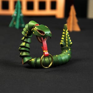 Op de afbeelding: Een groene leren slangen sleutelhanger met een rode tong en een gouden ring. De slang is opgerold in een cirkel met zijn kop naar voren gericht.