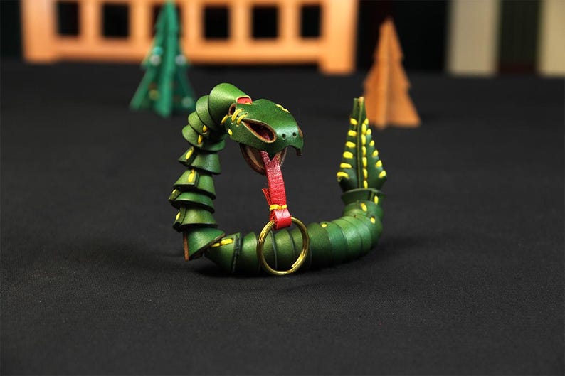 Op de afbeelding: Een groene leren slangen sleutelhanger met een rode tong en een gouden ring. De slang is opgerold in een cirkel met zijn kop naar voren gericht.