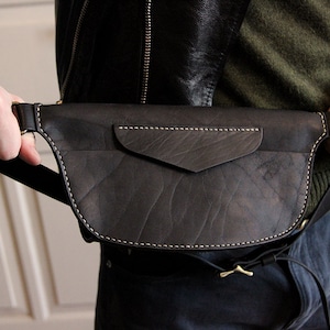 May include: A black leather fanny pack with a flap pocket and a white stitch detail. The bag is worn around the waist of a person.