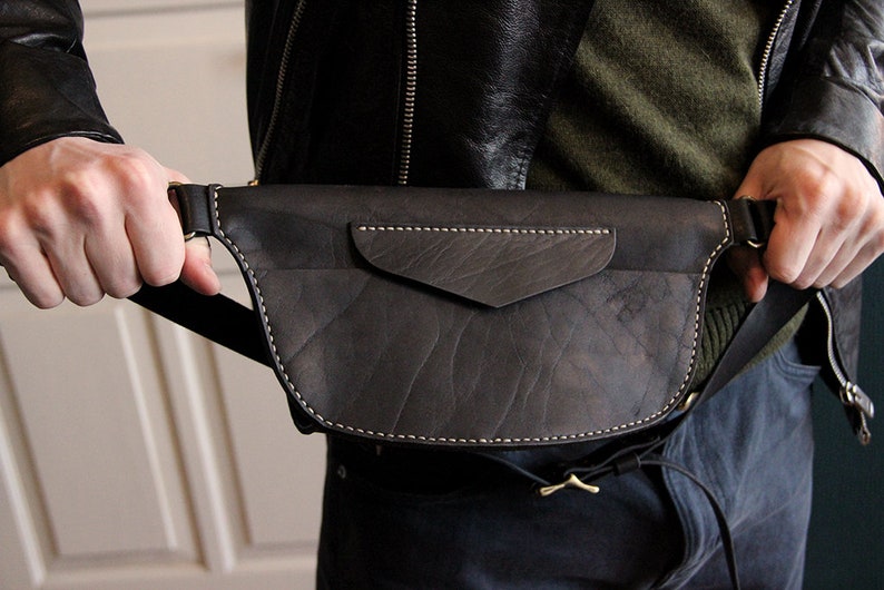 May include: A black leather fanny pack with a flap pocket and a white stitch detail. The bag is worn around the waist of a person.