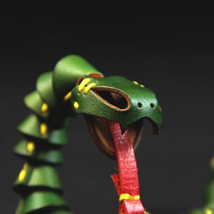Op de afbeelding: Een groene leren slangen-sculptuur met gele stiksels en een rode tong. De slang is opgerold en heeft een gedetailleerd hoofd.