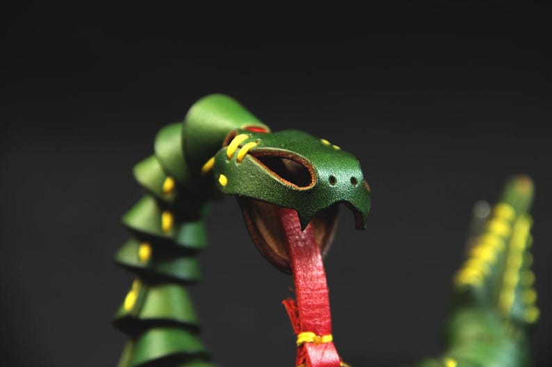 Op de afbeelding: Een groene leren slangen-sculptuur met gele stiksels en een rode tong. De slang is opgerold en heeft een gedetailleerd hoofd.