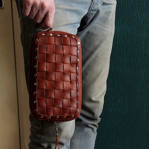 May include: A brown leather clutch bag with a woven design and silver studs. The bag has a zipper closure and a small tassel.