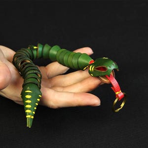 Op de afbeelding: Een groene en gele leren slangen sleutelhanger met een rode tong en een gouden sleutelring.