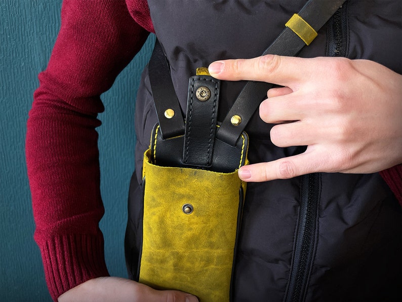 May include: A yellow leather phone case with a black leather strap and gold accents. The case is worn on a person's body.