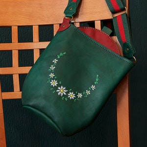 May include: A green leather shoulder bag with a red lining and a red and white striped strap. The bag has a white daisy embroidery design.