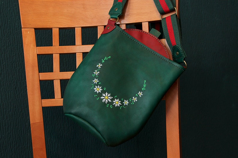 May include: A green leather shoulder bag with a red lining and a red and white striped strap. The bag has a white daisy embroidery design.