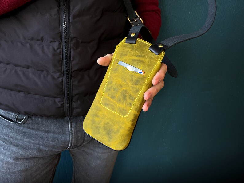 May include: A yellow leather phone case with a black strap. The case has a card slot and is worn over the shoulder.