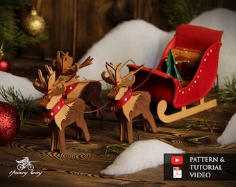 May include: Two wooden reindeer pulling a red felt sleigh with a green Christmas tree inside. The reindeer are decorated with red and gold accents. The sleigh is decorated with gold glitter. The scene is set on a wooden surface with white faux snow.
