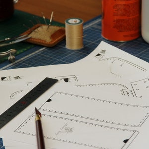 May include: A close-up of a leather crafting project with a ruler, a pen, and several paper patterns. The patterns include a rectangle with a dotted line border and a curved shape with a dotted line border.