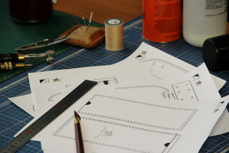 May include: A close-up of a leather crafting project with a ruler, a pen, and several paper patterns. The patterns include a rectangle with a dotted line border and a curved shape with a dotted line border.