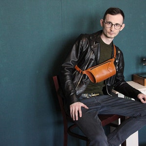 May include: A person wearing a black leather jacket, a green shirt, and blue jeans is sitting on a wooden chair. They are wearing a brown leather fanny pack worn across their chest. The person is wearing glasses and has short brown hair. The background is a teal wall.