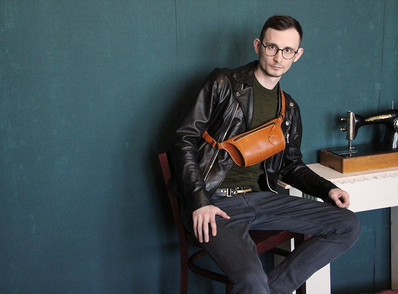 May include: A person wearing a black leather jacket, a green shirt, and blue jeans is sitting on a wooden chair. They are wearing a brown leather fanny pack worn across their chest. The person is wearing glasses and has short brown hair. The background is a teal wall.