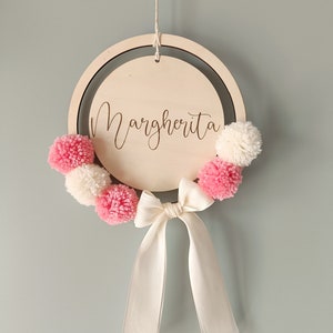 Può includere: Un cerchio di legno con il nome "Margherita" inciso su di esso. Il cerchio è decorato con pompon rosa e bianchi e un nastro di raso bianco.