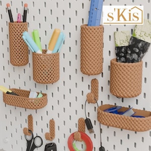 Può includere: Un pannello forato bianco con cesti intrecciati marroni e ganci. I cesti sono pieni di vari oggetti, tra cui penne, matite, pennelli e nastro adesivo. Il pannello forato ha il testo 's'Kis' nell'angolo in alto a destra.