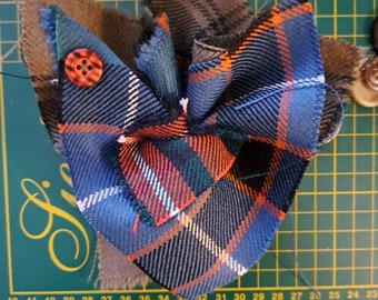 Può includere: Un fiocco di stoffa a quadri blu, arancio e verde con un retro in tessuto marrone e un bottone arancione al centro.