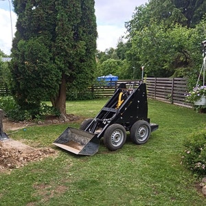 Op de afbeelding: Een zwarte minigraafmachine met een gele bak staat op een gazon. De graafmachine heeft twee grote achterwielen en één kleiner voorwiel. Er is een schop en een hoop aarde op de voorgrond.