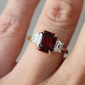 以下が含まれることがあります： 中央に大きな長方形の濃い赤色の宝石をあしらい、両側に2つの小さな長方形の透明な宝石を配したゴールドリング。 マニキュアされた爪のある手にリングがはめられています。