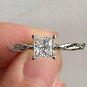 Puede incluir: Un anillo de compromiso plateado con un diamante de talla cuadrada. La banda presenta un diseño de cuerda trenzada. El anillo se sostiene sobre un fondo suave y neutro.