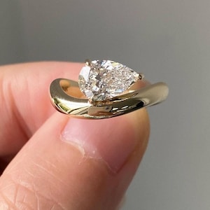 May include: A gold ring featuring a pear-shaped diamond set in a distinctive, curved band.
