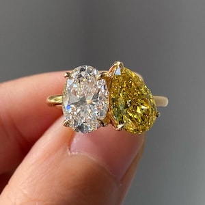 以下が含まれることがあります： 2つの楕円形のダイヤモンドがセットされた金の指輪。1つのダイヤモンドは透明で、もう1つは黄色です。