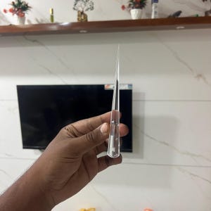 Könnte beinhalten: Ein klares, spitzes Glasobjekt wird in einer Hand gehalten. Das Objekt ist lang und schlank, mit einer scharfen Spitze oben. Das Glas scheint transparent zu sein. Das Objekt wird vor einem Fernseher und einem Regal mit Dekorationen gehalten.