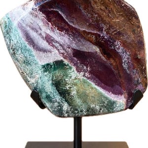 Pode incluir: Uma placa irregular e polida de mineral fluorita com faixas vibrantes de roxo profundo, verde floresta e marrom terroso, exibida na vertical sobre um suporte de metal preto com base quadrada e dois braços de suporte curvos.