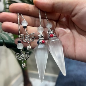 Puede incluir: Dos péndulos de cristal blanco translúcido con punta, suspendidos de cadenas plateadas. Cada péndulo presenta una tapa metálica decorativa con pequeños acentos rojos y azules, rematada con una cuenta blanca redonda y un dije de loto.