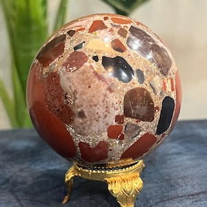 May include: A polished spherical stone featuring a brecciated pattern with a mix of red, brown, black, and cream-coloured mineral fragments embedded in a matrix. The sphere rests on a small, ornate, gold-coloured metal tripod display stand.