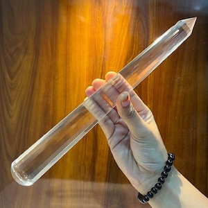 Puede incluir: Una varita de cristal transparente y alargada, con una punta facetada. La varita se sostiene en una mano, y una pulsera de cuentas negras es visible en la muñeca. El fondo es de madera.