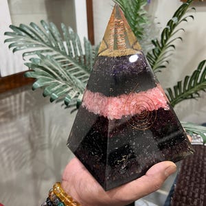 Puede incluir: Una pir&aacute;mide transparente hecha de cristales en capas. La pir&aacute;mide est&aacute; hecha de amatista, cuarzo rosa y turmalina negra. Una bobina de alambre de cobre est&aacute; incrustada en la pir&aacute;mide.