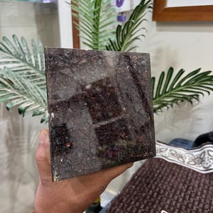 Puede incluir: Una pieza cuadrada de obsidiana marr&oacute;n oscuro, negro y rojo con una superficie rugosa y texturizada. La obsidiana est&aacute; incrustada con peque&ntilde;os trozos de metal brillantes.