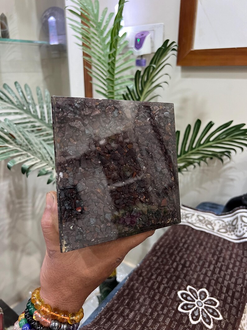 Puede incluir: Una pieza cuadrada de obsidiana marr&oacute;n oscuro, negro y rojo con una superficie rugosa y texturizada. La obsidiana est&aacute; incrustada con peque&ntilde;os trozos de metal brillantes.